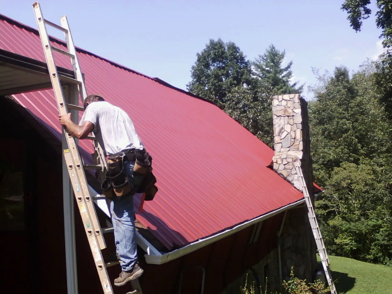 Roofer on a ladder beside a red metal roof and stone chimney — Soffit & Fascia Repair by Roofing Brothers in Mentor